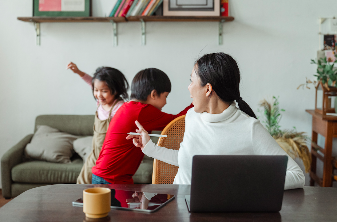 mom at home working with laptop and two kids behind her running around